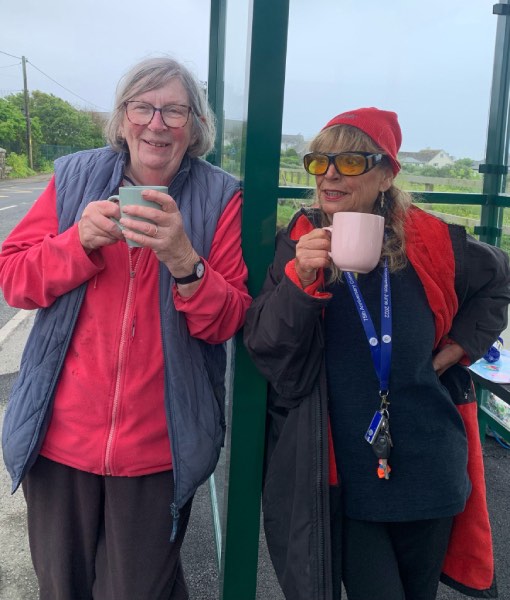 Two women enjoy a cup of tea and a chat leaning against a bus shelter in Sennen