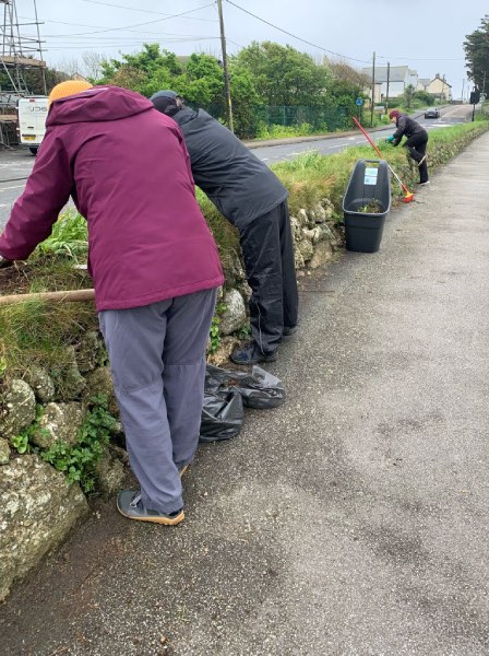 Three volunteers bend over to work on a cornish hedge in Sennen, the wheelbarrow, broom and bin bags that they are using are nearby.